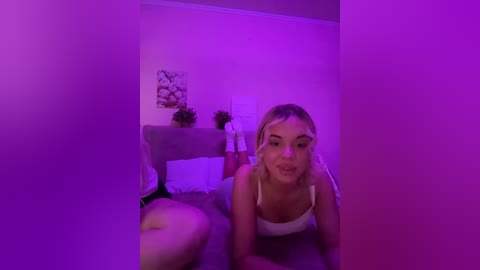 Video of a blonde woman in a white tank top, leaning forward with a playful expression, in a dimly lit room with purple lighting, surrounded by plants and framed art on a wooden desk.