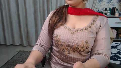 Video of a South Asian woman with light skin, wearing a beige kurta with intricate embroidery, a red scarf, and a floral-patterned dupatta. Background includes beige curtains, a white dresser with cosmetics, and a blue-patterned bedspread.