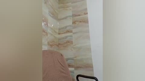 A video shows a beige tiled bathroom wall with a light pink towel draped over a black chair, suggesting a recently used shower. The tiles have a marbled pattern in beige and light brown hues.