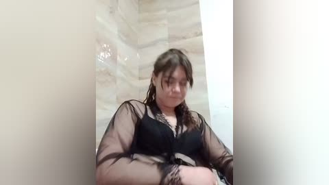 Video of a young Caucasian woman with fair skin, brown hair, and a slender build, wearing a sheer black lace bra and matching robe, sitting in a tiled bathroom.