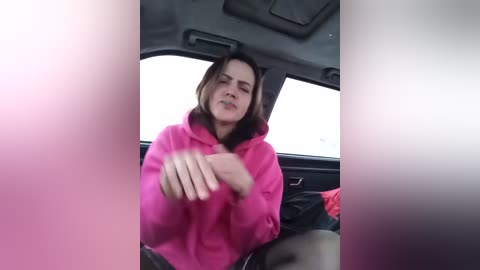 A candid video captures a woman in a bright pink hoodie, sitting in a car, with blurred background. She appears relaxed, with a slight smile.