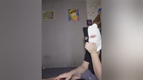 Video of a person wearing a black mask and white towel over head, standing in a dimly lit room with gray walls and colorful posters.