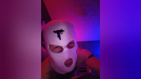 Video of a person wearing a white balaclava with a black anarchist symbol, lit by purple and red neon lights, creating a stark, moody atmosphere.
