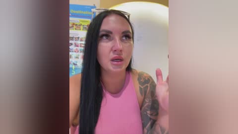 Video of a tattooed woman with long black hair, wearing a pink top, and sunglasses, speaking animatedly in a classroom with colorful posters and a large globe.