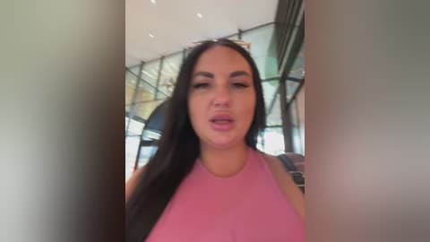 A video of a young woman with long black hair, wearing a pink tank top, standing in a modern, glass-encased building with large windows and metal frames.