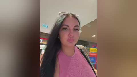 Video of a young woman with long black hair, wearing a pink tank top, sunglasses on her head, and a black jacket. She stands in a brightly lit, modern indoor space with colorful shelves in the background.