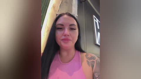 Video of a smiling, light-skinned woman with long black hair and pink lips, wearing a sheer pink top, standing in a dimly lit, rustic room with exposed concrete walls and a partially visible neon sign.