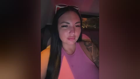 Video of a young woman with long black hair, wearing a sleeveless pink top, and sunglasses on her head. She has a tattoo on her arm and a neutral expression. Dimly lit room with red and orange hues.