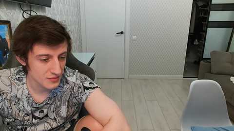 Video of a young Caucasian man with short brown hair, wearing a graphic t-shirt, sitting at a glass table in a modern living room with light wood flooring and white patterned wallpaper.
