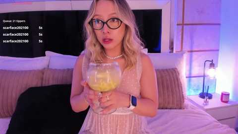 Video of a blonde woman in glasses, wearing a white sleeveless dress, holding a glass of orange liquid, on a bed with white sheets and pillows, in a dimly lit room.