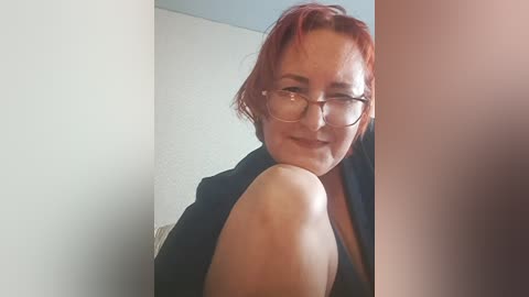 A video of a middle-aged woman with short, dyed red hair, glasses, and a black top, smiling while leaning against a white wall.