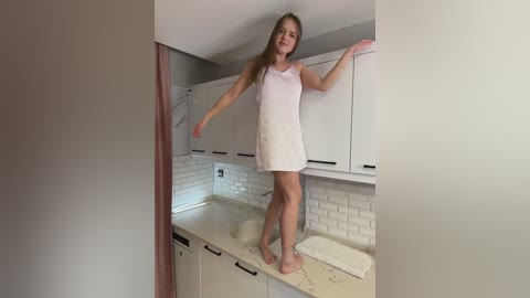 Video of a young, fair-skinned, long-haired girl in a pink sleeveless dress, barefoot, balancing on a kitchen counter. White cabinets, subway tiles, and a cream towel are visible.