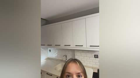 Video of a modern kitchen with white cabinets, white subway tiles, and a white countertop. A blonde woman with a short hairstyle is partially visible, peering through the kitchen door.