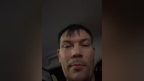 A close-up video of a middle-aged man with short black hair and a serious expression, taken from a low angle, showing his upper torso. The background is dimly lit and out of focus.