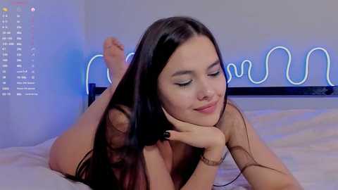 A video of a nude, fair-skinned woman with long black hair lying on her stomach on a bed. She wears minimal jewelry and has a serene expression. The background features a glowing neon sign.