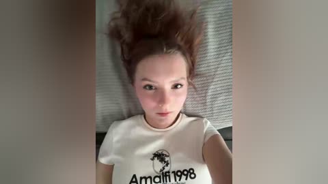 Video of a young woman with fair skin, large eyes, and dark brown hair, lying on her back, wearing a white t-shirt with \"Amalfi 1988\" text and a globe design. Background shows a textured, light grey blanket.