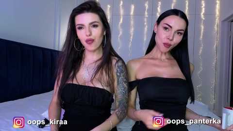 Video of two women with fair skin and long dark hair, wearing strapless black dresses, standing in a bedroom with white curtains and a dark headboard.