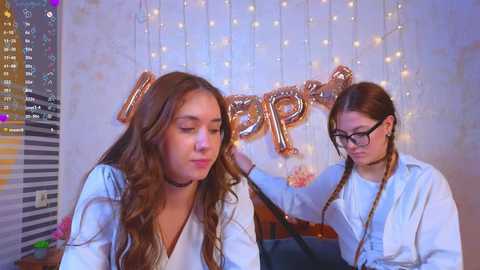 Video of two young women with long brown hair, one with glasses, wearing white shirts and chokers, blowing up metallic \"21\" balloons against a glittery backdrop.
