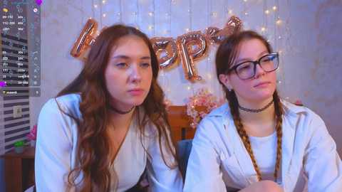 Video of two young women with long hair, wearing white tops, sitting close. Background features \"Happy Birthday\" balloons and string lights.