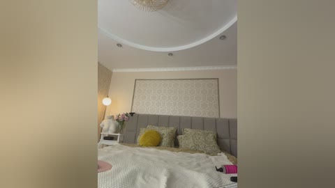 Video of a modern, cozy bedroom featuring a tufted grey headboard, floral pillows, a white knitted blanket, a floral-patterned wall, and a spherical lamp on a side table.