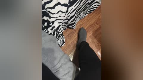 Video of a person with light skin wearing a gray sweater and black pants, sitting on a wooden chair with a zebra-patterned cushion.