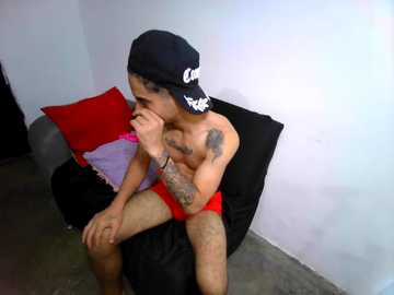 Video of a shirtless, muscular, tanned man with a black cap and red briefs, sitting on a black beanbag chair, leaning on his left arm, in a simple, dimly lit room with a white wall and red pillow.