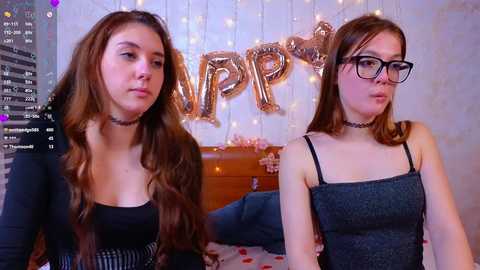 Video of two young women with long wavy hair, wearing black chokers and dresses, in a room with a \"Happy Birthday\" banner and twinkling fairy lights.