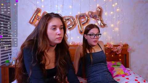 Video of two young women with long hair, one in glasses, wearing dark clothing, sitting on a bed with pink sheets, surrounded by gold \"BIRTHDAY\" balloons and fairy lights.