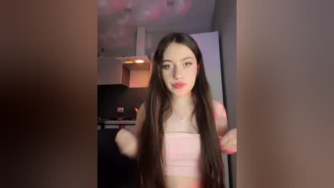 A video of a young woman with long, straight brown hair, fair skin, and wearing a pink crop top. She stands in a modern kitchen with a white ceiling and dark cabinets.