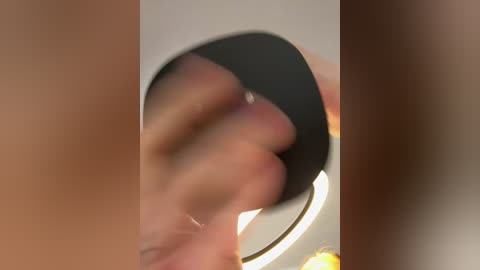 Video of a blurred, close-up hand holding a black object, with a blurred background featuring warm tones and a circular light source. The hand appears to be gripping the object firmly.