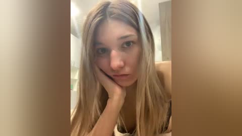 Video of a young woman with long, straight, light brown hair, resting her chin on her hand, wearing a beige top. The background is blurred, showing a neutral-colored room with white walls.