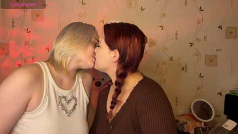 A video of two women kissing passionately, one with short platinum blonde hair, the other with long auburn hair, in a dimly lit room with butterfly wallpaper and a mirror.