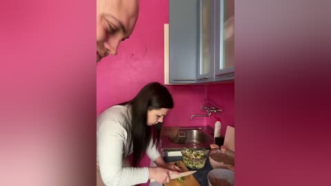 Video of a woman with long black hair in a white shirt chopping vegetables on a pink countertop in a small kitchen with blue cabinets and a stainless steel sink.