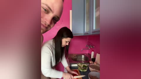 Video of a woman with long dark hair, wearing a white top, chopping vegetables in a pink kitchen with stainless steel appliances, and a man's face blurred in the background.