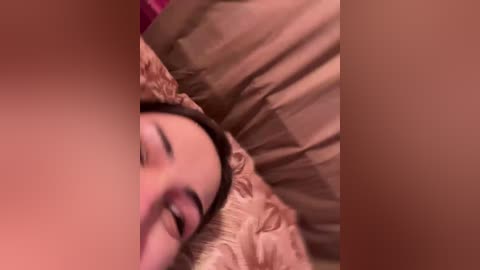 A blurry video of a woman lying on a bed, her head obscured by pillows. The background shows a beige bedspread and a pink wall, creating a cozy, intimate setting.
