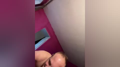 Video of a naked man with short, light brown hair, captured from a low angle, peering through a small rectangular window in a vibrant pink wall.