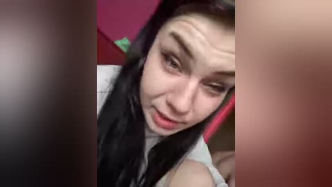 Video of an Asian woman with long black hair and fair skin, wearing a white top, looking down. Background includes a pink surface and blurred figures.