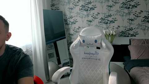 Video of a man in a gray shirt seated in a white gaming chair, with a large flat-screen TV, white bedding, and floral wallpaper in the background.