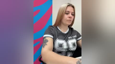 Video of a young woman with light skin and long blonde hair, wearing a gray \"Not 69\" T-shirt. She has a tattoo on her right arm. Background features a vertical blue and pink pattern.