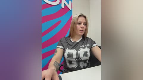 Video of a blonde woman in a New York Giants jersey, sitting at a table, with a colorful background featuring a blue and pink \"SUN\" sign.