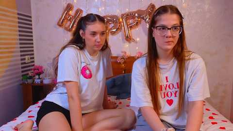 Video of two young women in white T-shirts with red heart print, sitting on a bed with heart-patterned sheets. One has long brown hair, the other glasses; background has gold \"Happy Birthday\" banner and floral wallpaper.