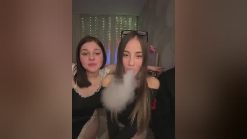 A video of two young women, one blowing a cloud of smoke, wearing black dresses, in a dimly lit room with a window and a pink wall.