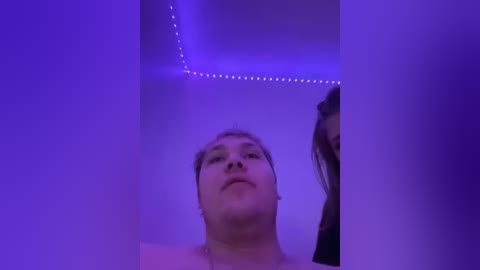 Video of a close-up, light-skinned, young man with short, light brown hair and a neutral expression. The background is purple with LED strip lights.