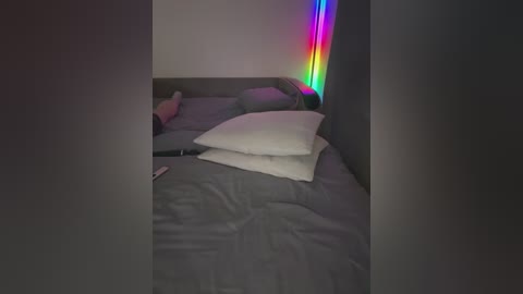 Video of a dimly lit bedroom with a gray bed, a white pillow, and a pink pillow, featuring a colorful LED strip on the wall, creating a vibrant backdrop.