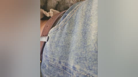Video of a close-up of a person's torso, showing a faded blue denim shirt with a white undershirt peeking out, and a brown belt. The background is blurred, featuring a textured, grayish fabric.