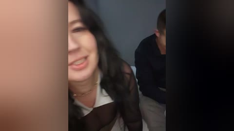 A video of a smiling woman with long black hair partially obscured by a dark pillar. A man with short brown hair sits nearby, wearing a dark shirt.