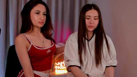 Video of two young Asian women with long black hair, wearing casual red tops and white shirts, sitting indoors.