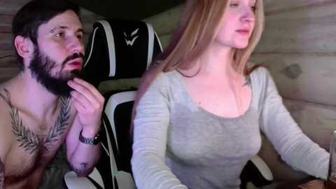 Video of a bearded man in a tribal tattoo shirt and a blonde woman in a low-cut gray top, seated in a black gaming chair with a \"W\" logo, in a dimly lit room with wooden walls.