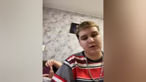 Video of a young, fair-skinned boy with short brown hair, wearing a striped red, white, and gray shirt, standing in a room with gray, textured wallpaper and a TV mounted on the wall.
