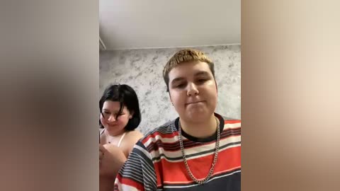 Video of a plus-size woman with fair skin and short brown hair in a striped shirt, standing behind a slender, dark-haired woman with glasses, both facing a white, floral-patterned wallpaper.